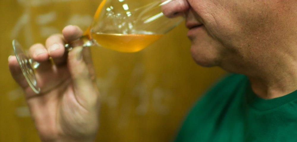 Armand in a green shirt closely examining a glass of beer in front of wooden foeders.