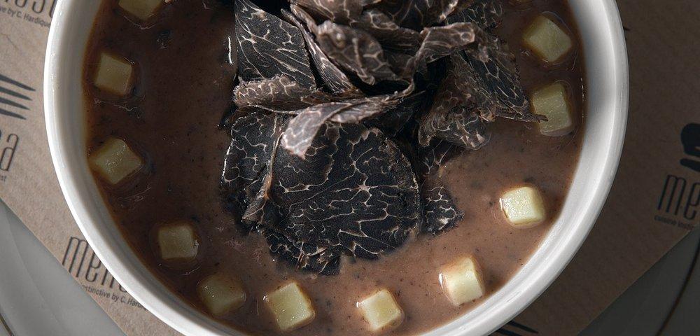 Vue du dessus d'un bol de velouté brun garni de lamelles de truffe noire et de petits cubes, posé sur une serviette en papier siglée Menssa.