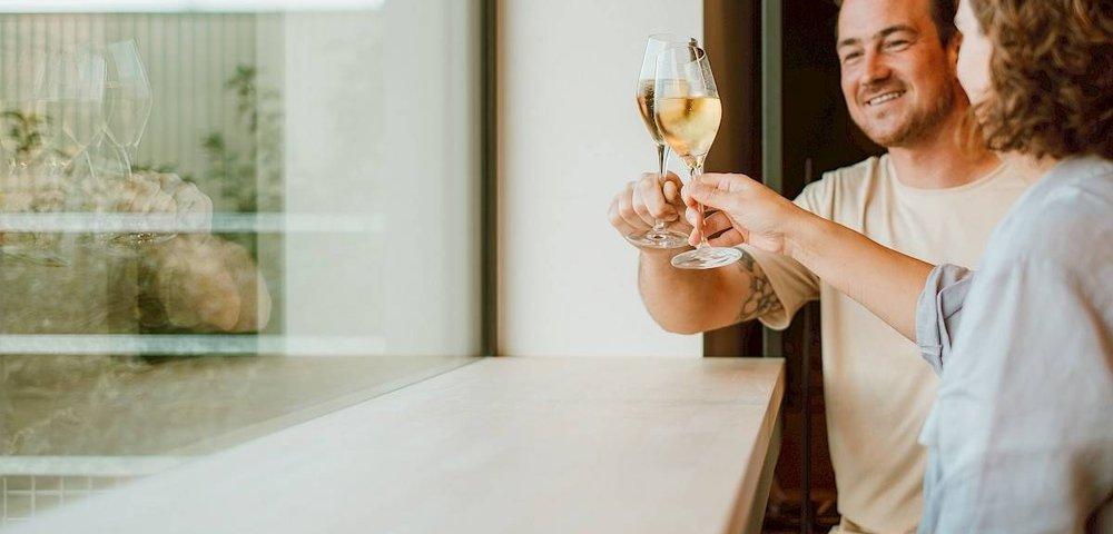 Twee mensen klinken met glazen witte wijn aan een houten bar bij een raam