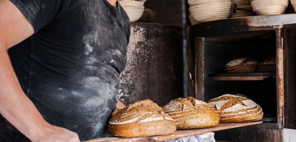 Bakker met bloemvlekken op zijn T-shirt haalt met een houten schep drie desembroden uit de oven, met rijsmandjes op de voorgrond