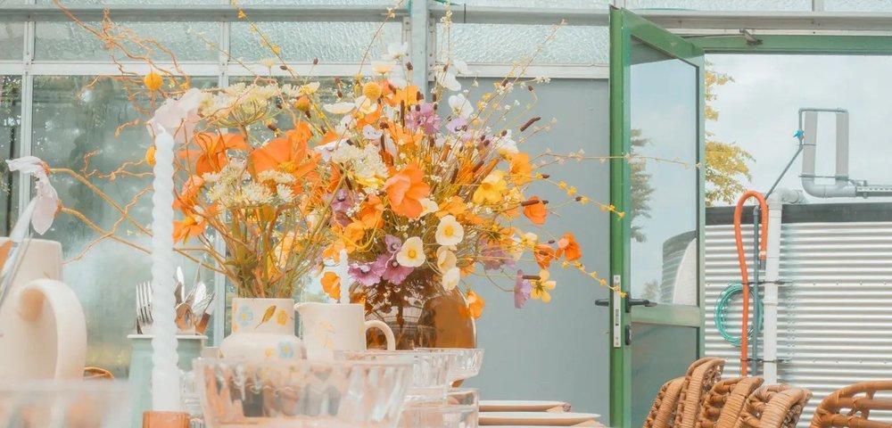 Lange houten tafel in de serre gedekt met borden, glazen en een boeket veldbloemen onder oranje hanglampen