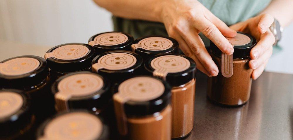 Mains fermant des pots de pâte à tartiner au chocolat à couvercle noir sur un plan de travail en inox.