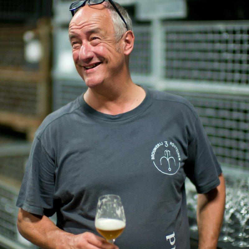 Portrait of Armand smiling in the warehouse, holding a glass of beer in a grey shirt.