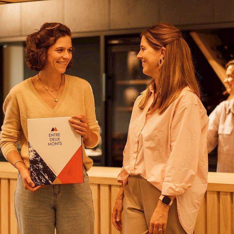 Twee vrouwen praten aan de toog, een van hen houdt een map met het logo Entre-Deux-Monts vast
