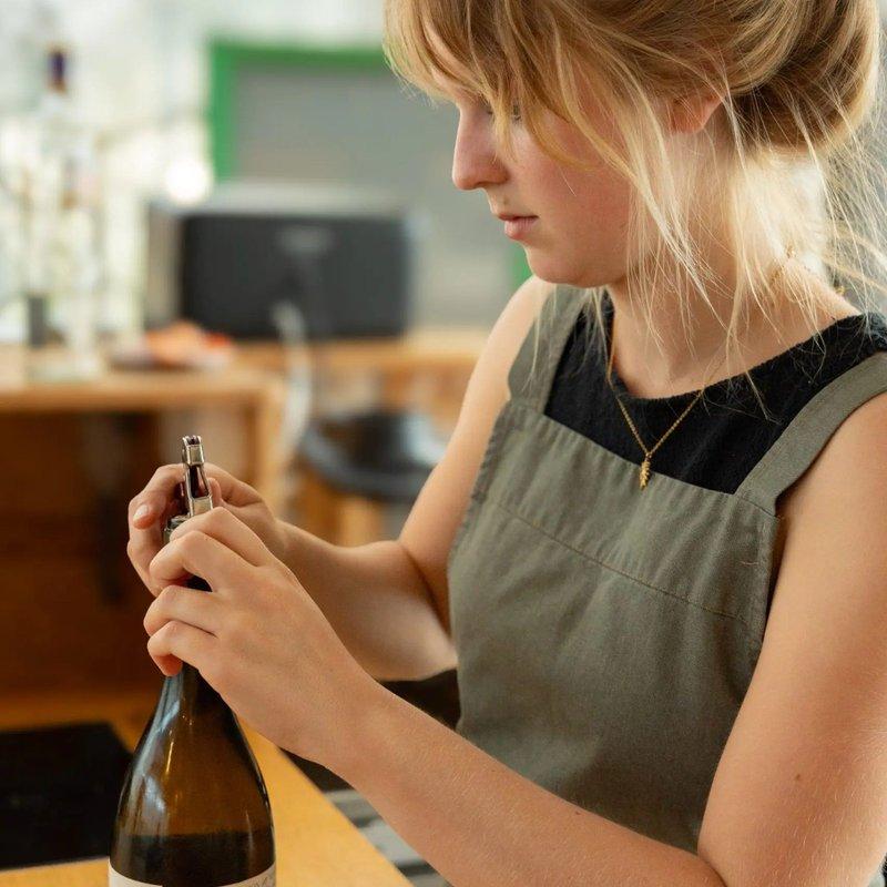 Vrouw met schort opent een fles wijn aan een houten toog