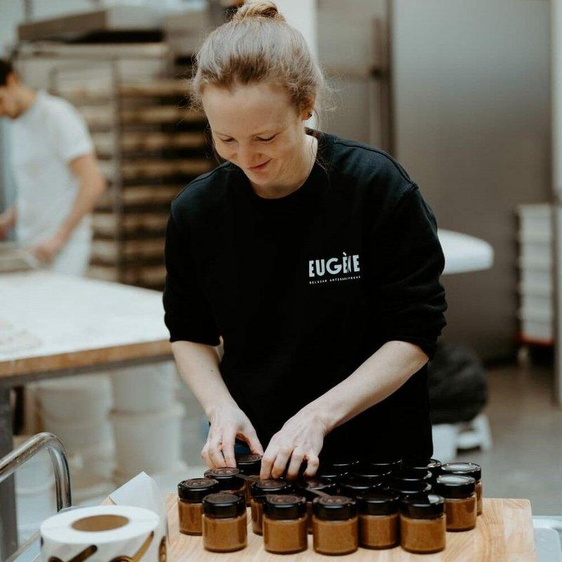 Une femme souriante en pull noir Eugène remplit des petits pots de pâte à tartiner dans un atelier.